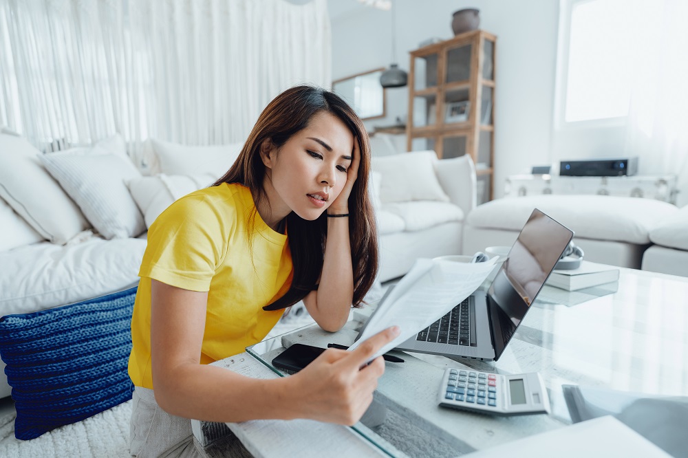 Une femme est assise par terre devant la table basse de son salon. Elle est penche sur un ordinateur portable et une calculatrice, des papiers  la main.