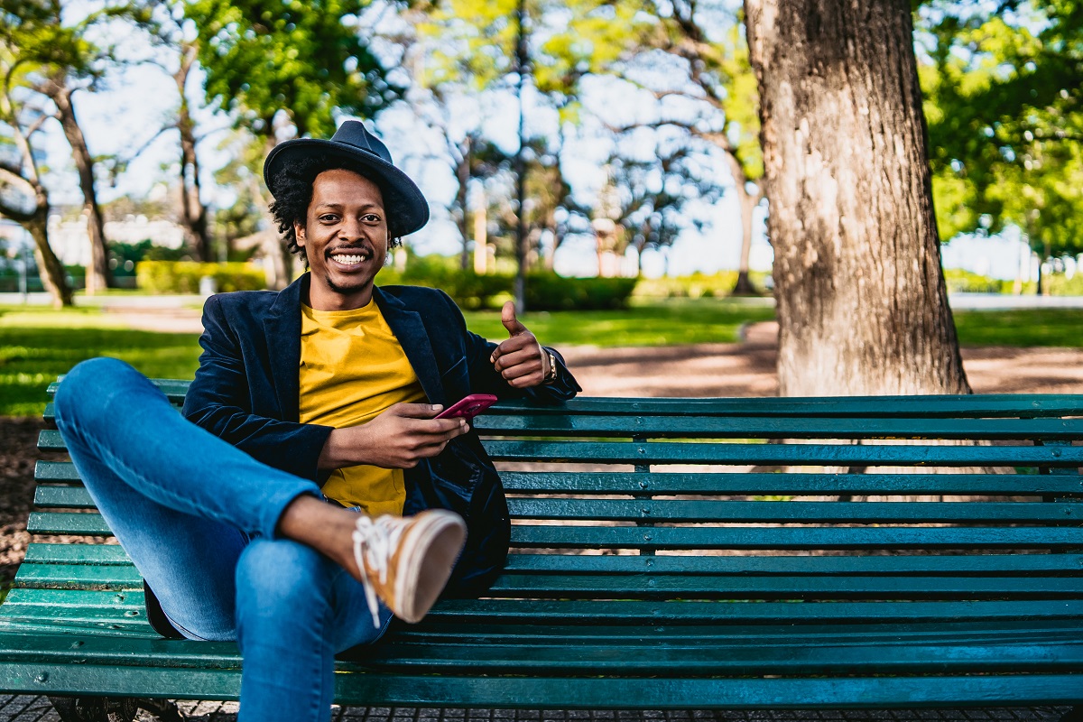 A young black man with a mobile phone, sitting on a bench in a park